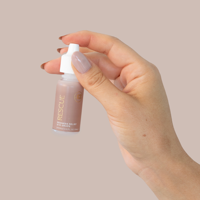 close up of a hand with tan nail polish holding an eye dropper bottle with tan label and gold text reading "rescue redness relief eye drops"