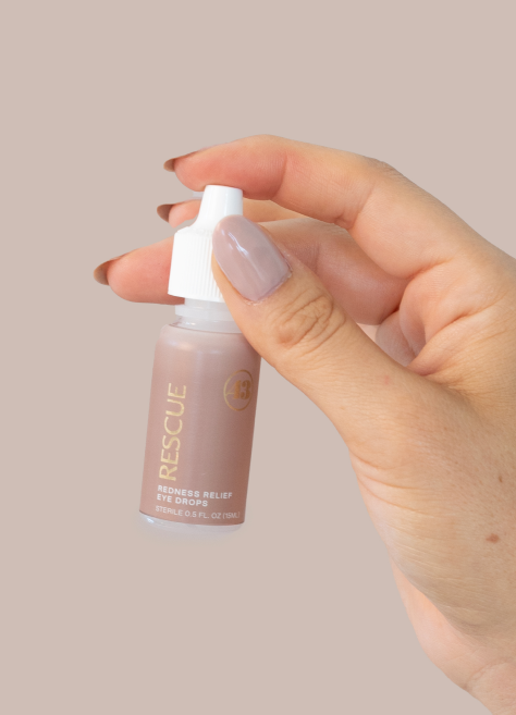 close up of a hand with tan nail polish holding an eye dropper bottle with tan label and gold text reading "rescue redness relief eye drops"