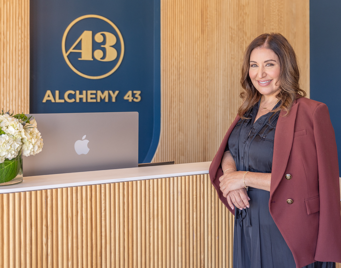 Alchemy 43 founder Nicci Levy wearing a navy dress and burgundy blazer posing in front of an Alchemy 43 front desk with computer monitor and white flowers. Alchemy 43 circle logo and gold "Alchemy 43 text" on the wall behind the desk